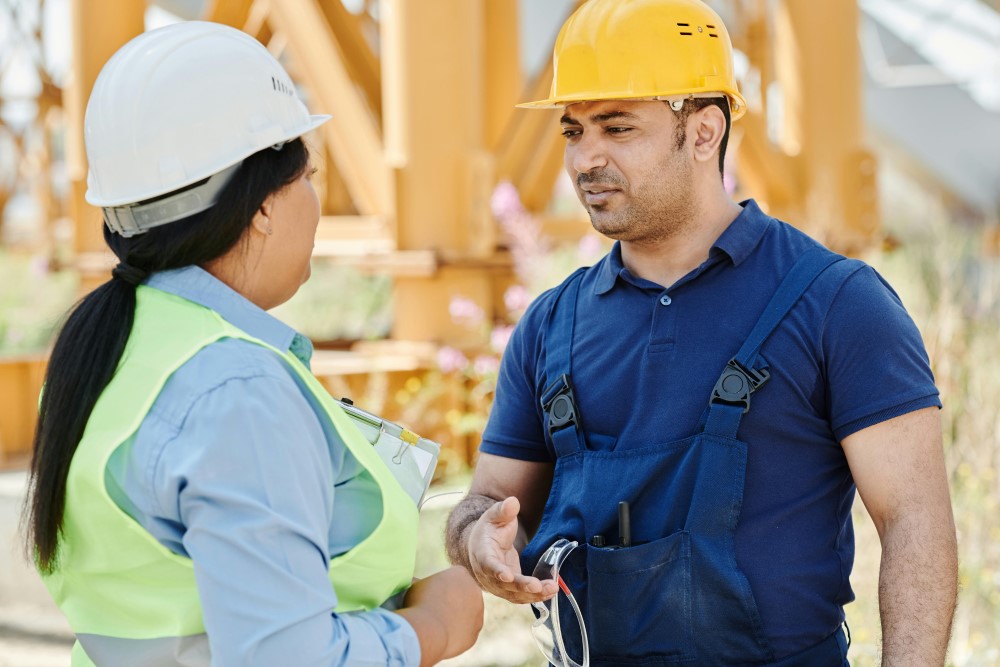 Two immigrant workers wearing safety equipment and hard hats talk on a construction site.