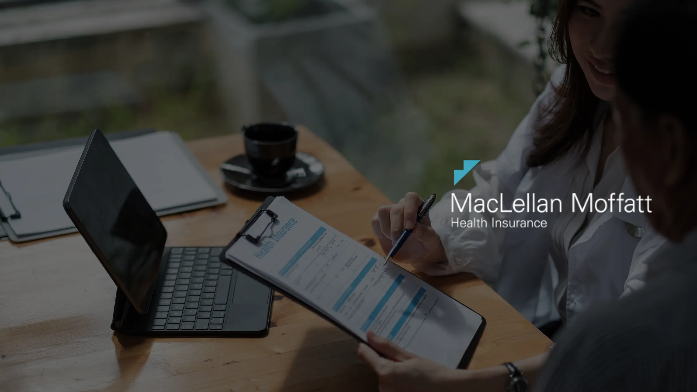 A hand holding a pen leans on a desk and fills out a form on a clipboard. There is a laptop and coffee mug on the desk. A MacLellan Moffatt Health Insurance logo is overlaid on the right of the image