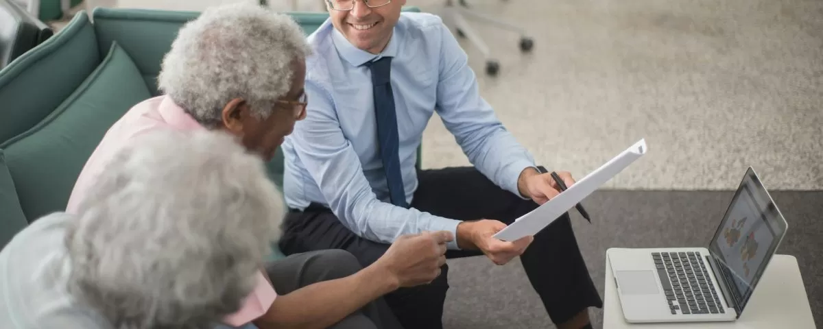 An insurance advisor holding paperwork sits on a sofa, smiling, next to an older couple. A laptop sits open on a low table in front of them