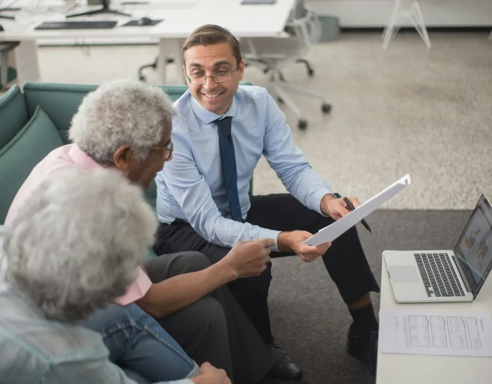 An insurance advisor holding paperwork sits on a sofa, smiling, next to an older couple. A laptop sits open on a low table in front of them