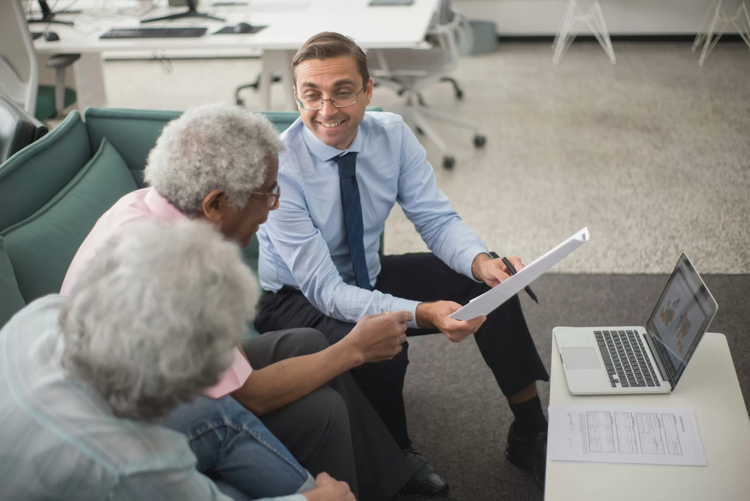 An insurance advisor holding paperwork sits on a sofa, smiling, next to an older couple. A laptop sits open on a low table in front of them