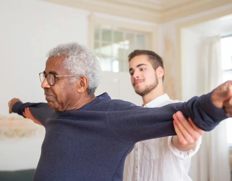 A senior man has his arms outstretched as a physiotherapist stands behind him, gently holding the mans arms back to assess his range