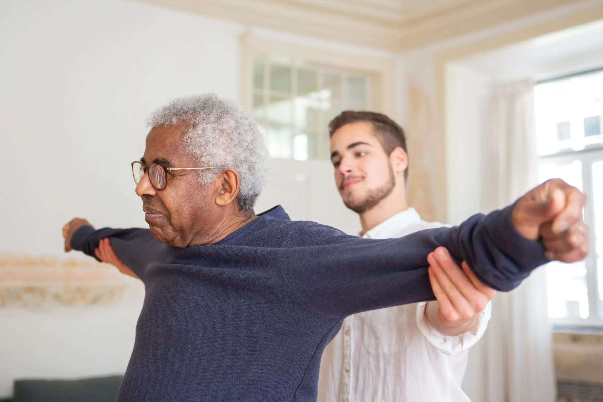 A senior man has his arms outstretched as a physiotherapist stands behind him, gently holding the mans arms back to assess his range