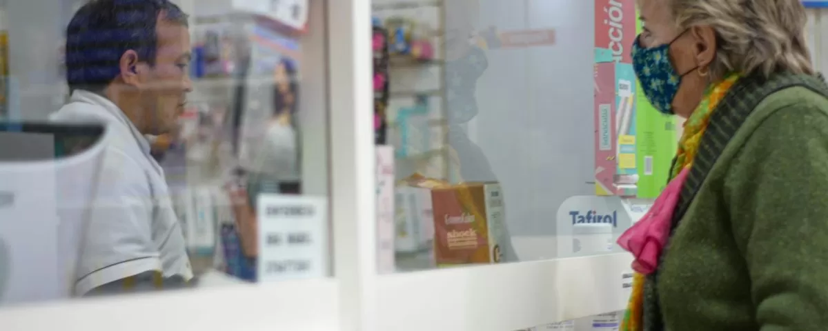 A female customer in her 50s stands at a pharmacy counter wearing a protective mask; a pharmacist stands behind plexiglass preparing a prescription