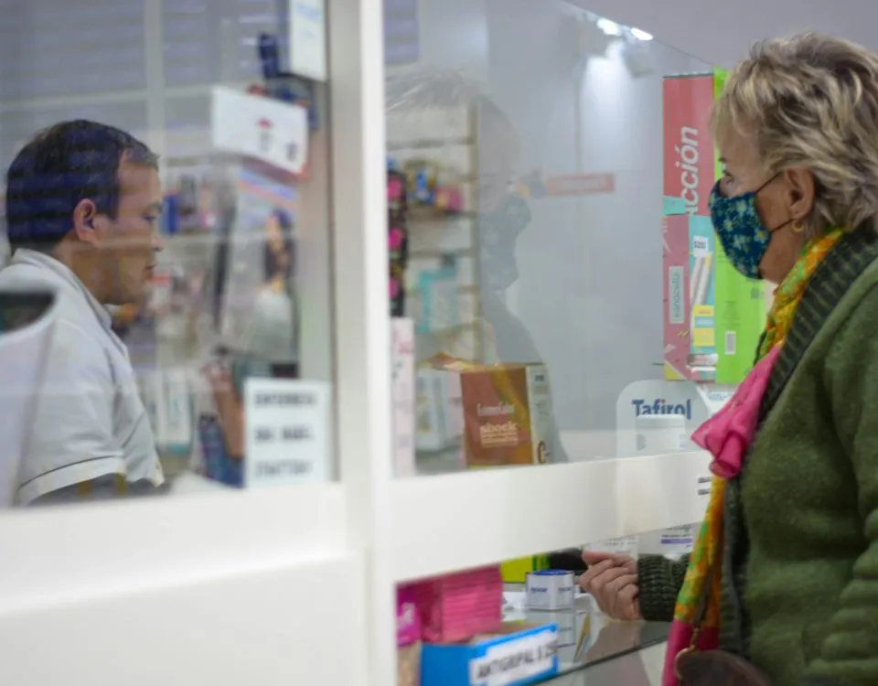 A female customer in her 50s stands at a pharmacy counter wearing a protective mask; a pharmacist stands behind plexiglass preparing a prescription