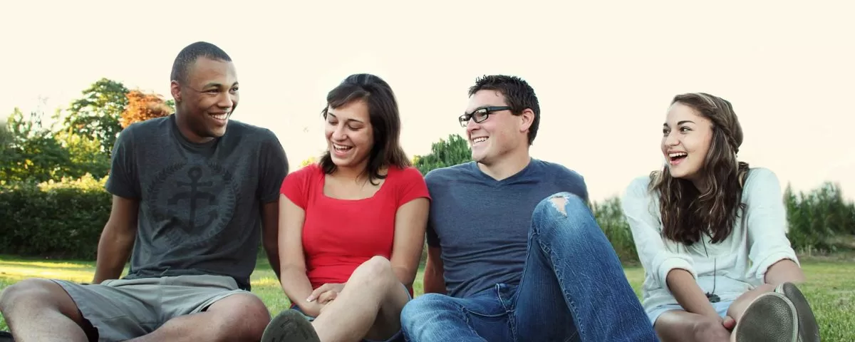 four youth sit in a row n a grassy field talking and laughing