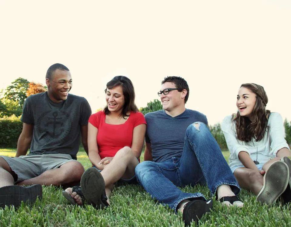 four youth sit in a row n a grassy field talking and laughing