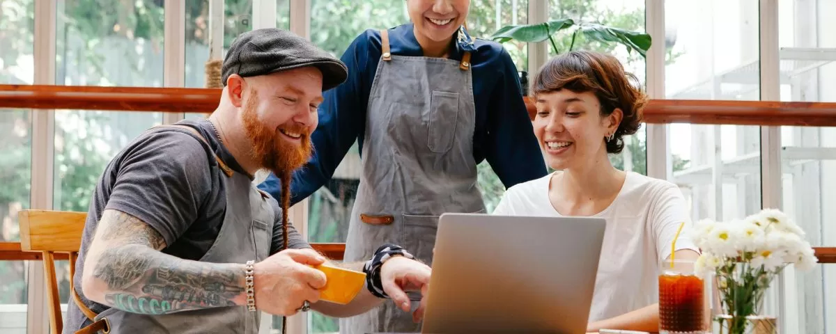 a 30-something man with a beard, baseball hat, and work apron sits at a table looking at a laptop and talking to a woman sitting next to him. A woman, also in a work apron, stands behind them also looking at the laptop and engaged in the conversation