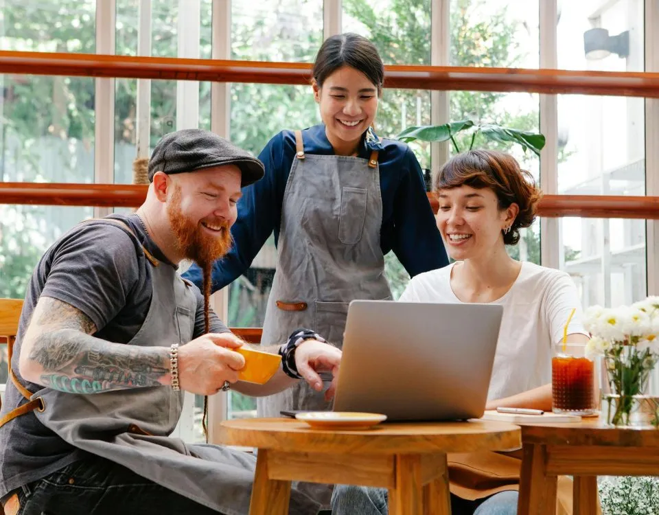a 30-something man with a beard, baseball hat, and work apron sits at a table looking at a laptop and talking to a woman sitting next to him. A woman, also in a work apron, stands behind them also looking at the laptop and engaged in the conversation