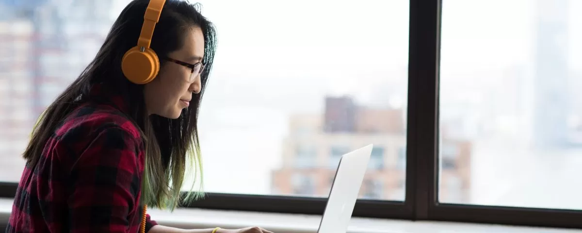 young Asian woman wearing yellow headphones sits at a table using a laptop