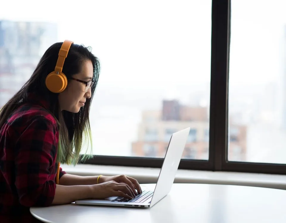 young Asian woman wearing yellow headphones sits at a table using a laptop