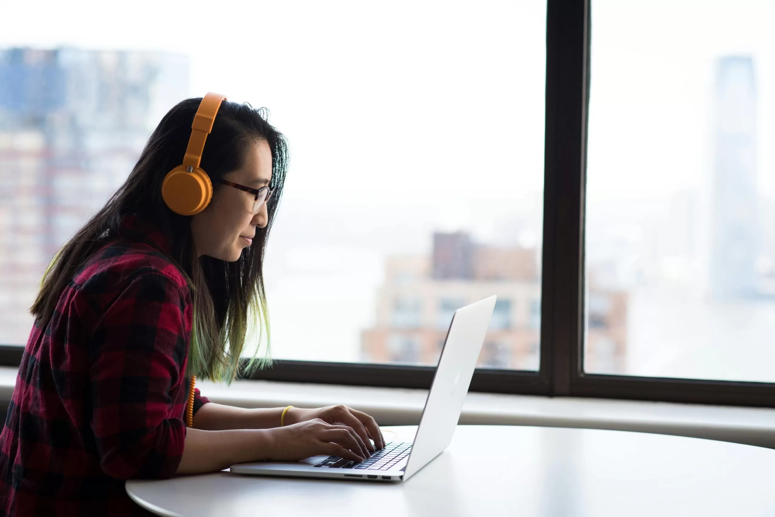 young Asian woman wearing yellow headphones sits at a table using a laptop