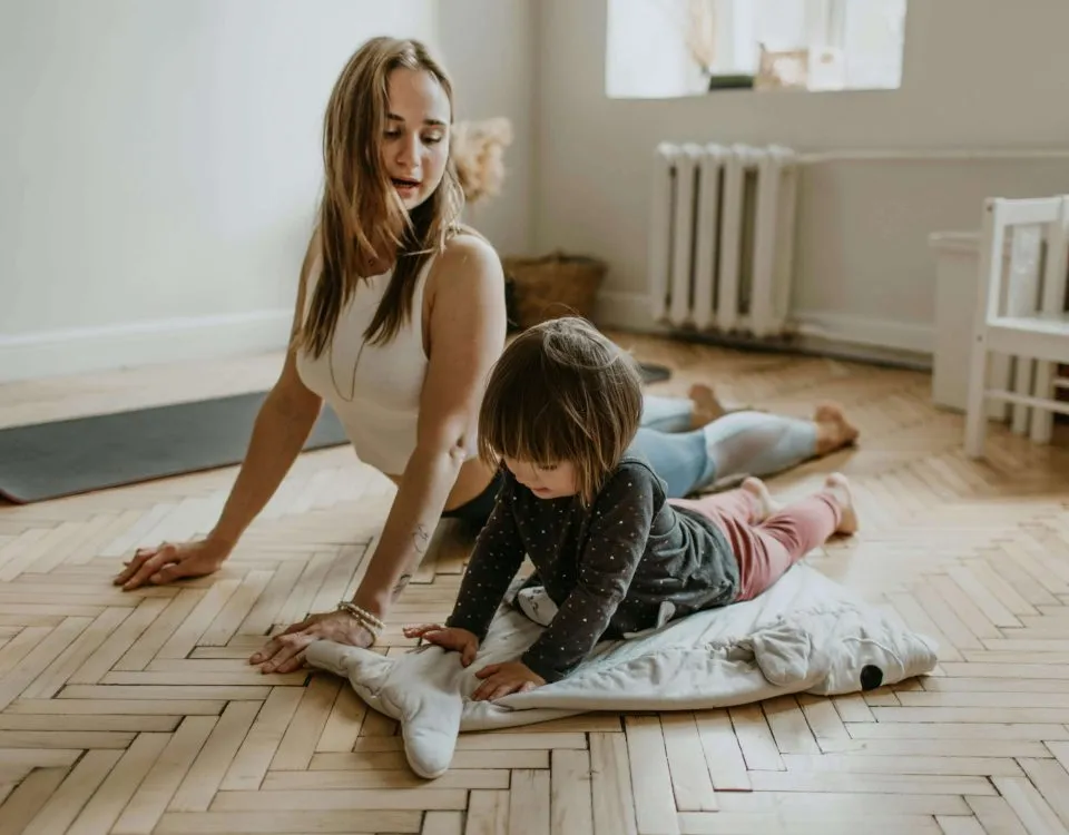 young woman in workout gear does a yoga pose on the floor while her toddler tries to mimic next to her