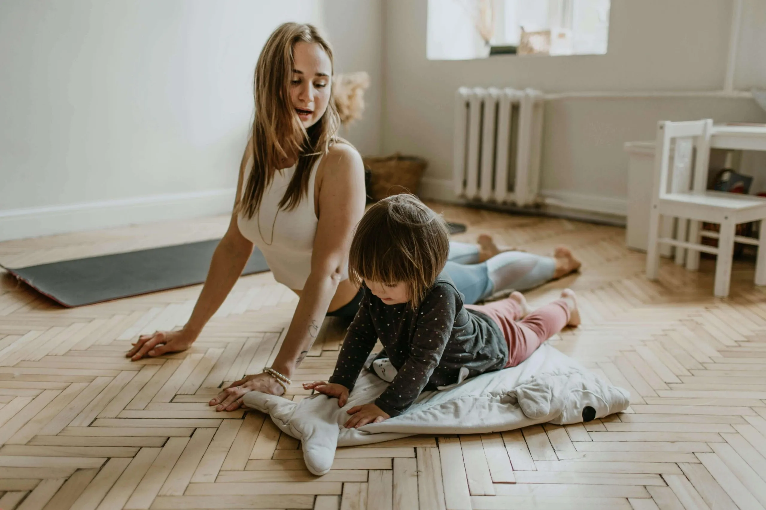 young woman in workout gear does a yoga pose on the floor while her toddler tries to mimic next to her