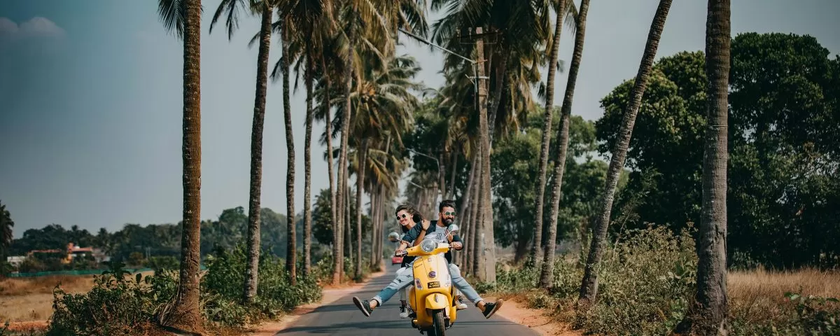 A man drives a yellow scooter toward the camera, on a road lined with palm trees. A woman sits behind him.