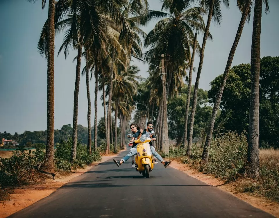 A man drives a yellow scooter toward the camera, on a road lined with palm trees. A woman sits behind him.