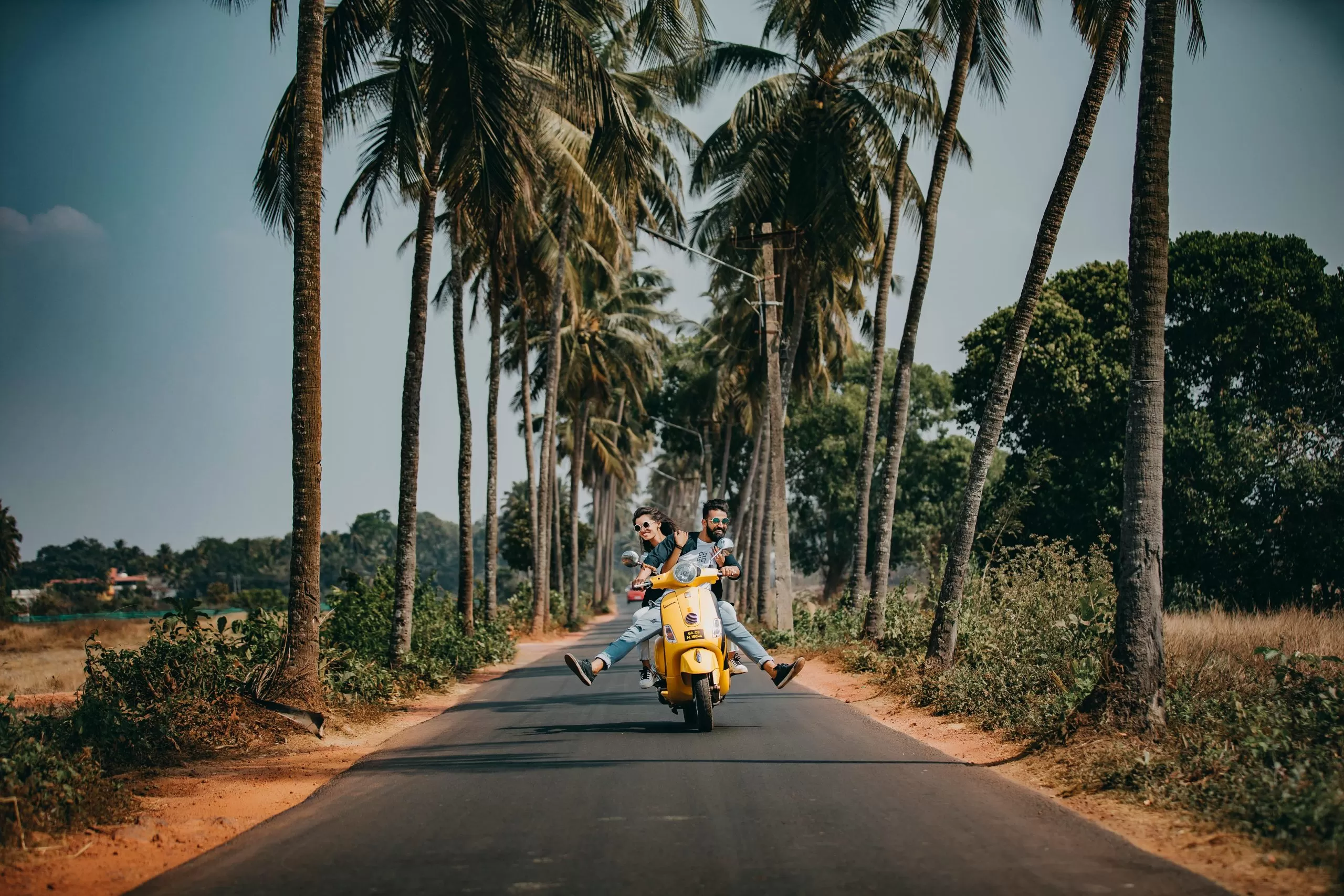 A man drives a yellow scooter toward the camera, on a road lined with palm trees. A woman sits behind him.