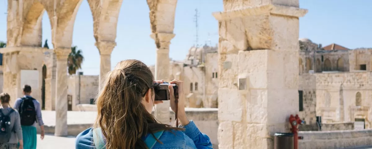 A young girl wearing a backpack is seen from behind taking a photo of an ancient landmark