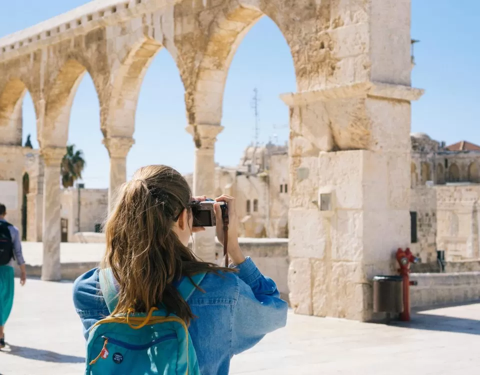 A young girl wearing a backpack is seen from behind taking a photo of an ancient landmark