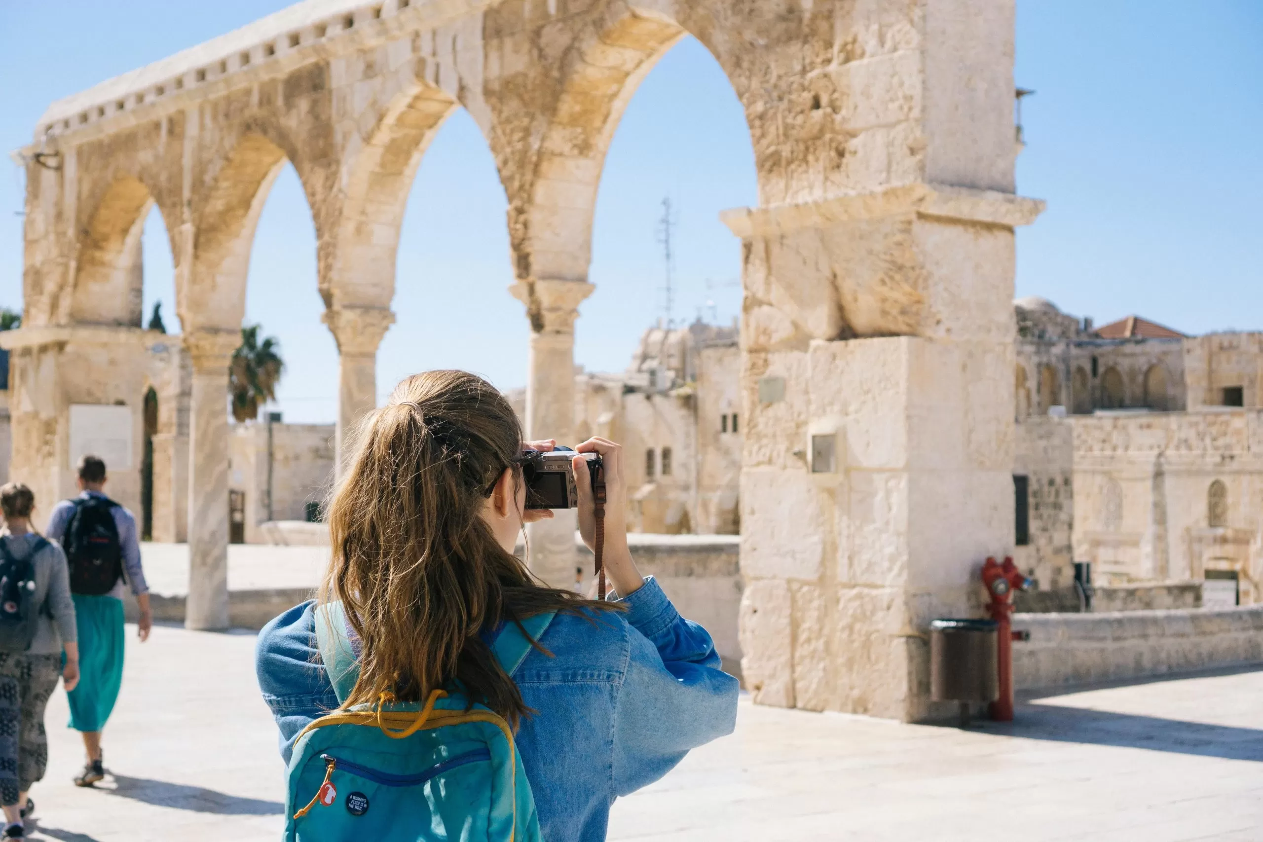 A young girl wearing a backpack is seen from behind taking a photo of an ancient landmark