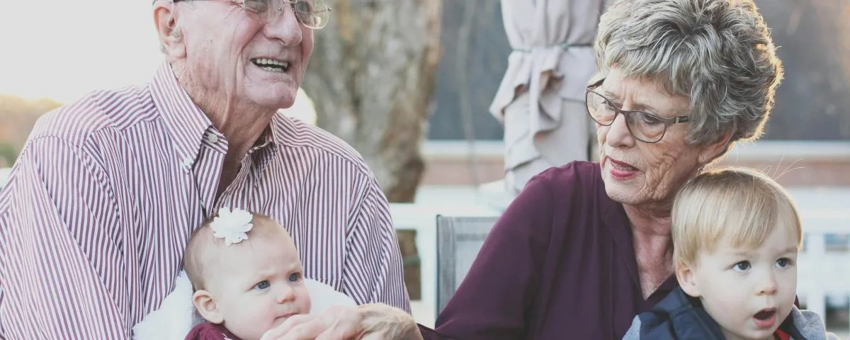 Grandparents in their 70s sit side by side, each with a grandbaby in their lap.