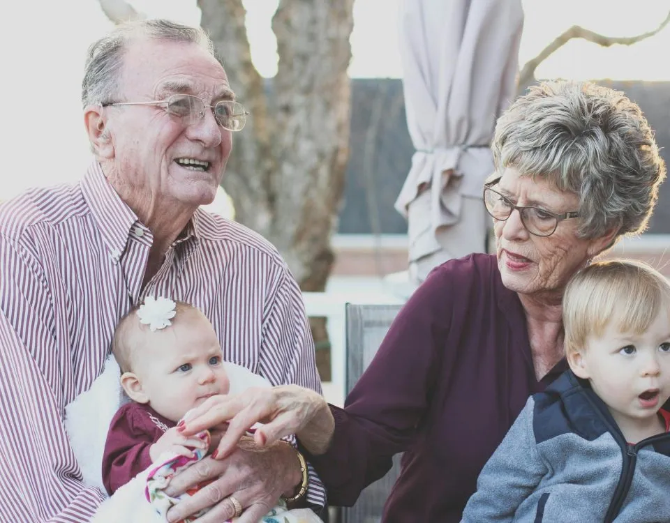 Grandparents in their 70s sit side by side, each with a grandbaby in their lap.