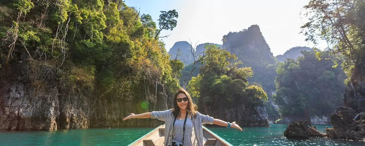 A woman sits cross-legged on a wooden dock with her arms outstretched, indicating the clear green water and tropical surroundings