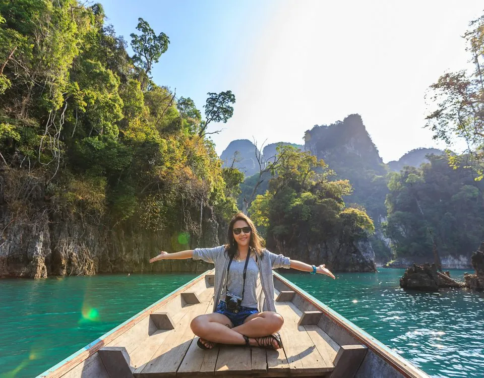 A woman sits cross-legged on a wooden dock with her arms outstretched, indicating the clear green water and tropical surroundings