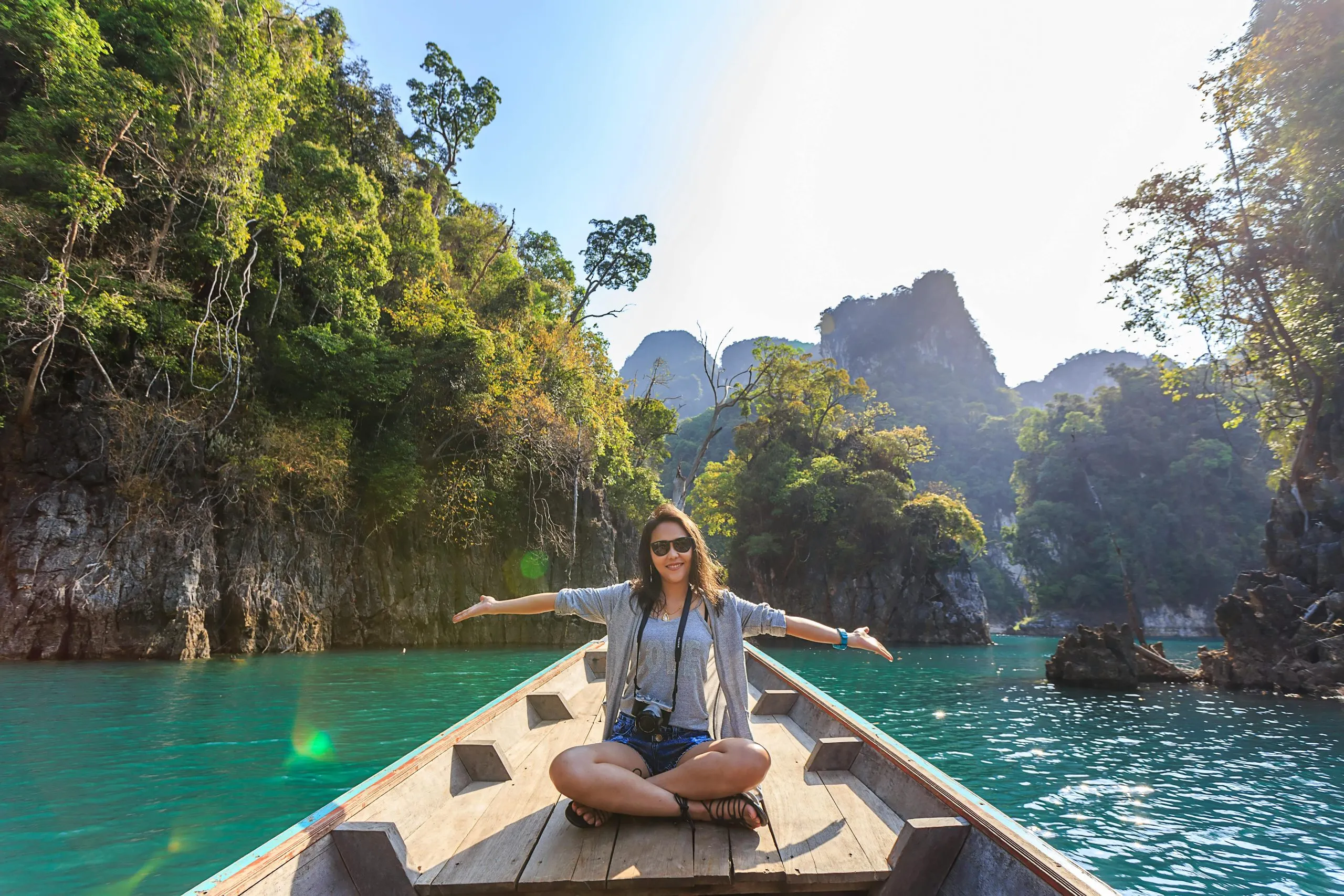 A woman sits cross-legged on a wooden dock with her arms outstretched, indicating the clear green water and tropical surroundings