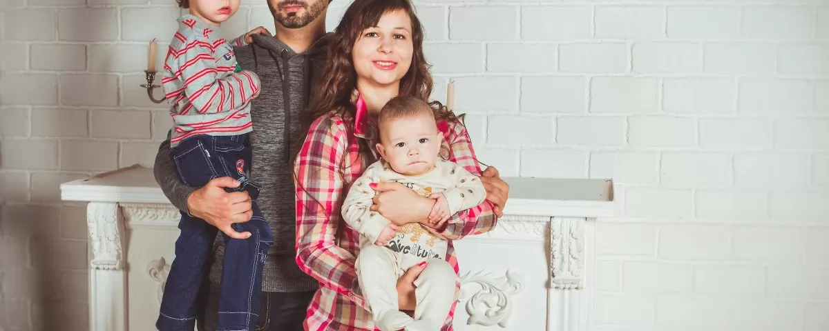A family poses in fron of a fireplace: 30-something dad holds a toddler and 30-something mom holds a baby in her arms.