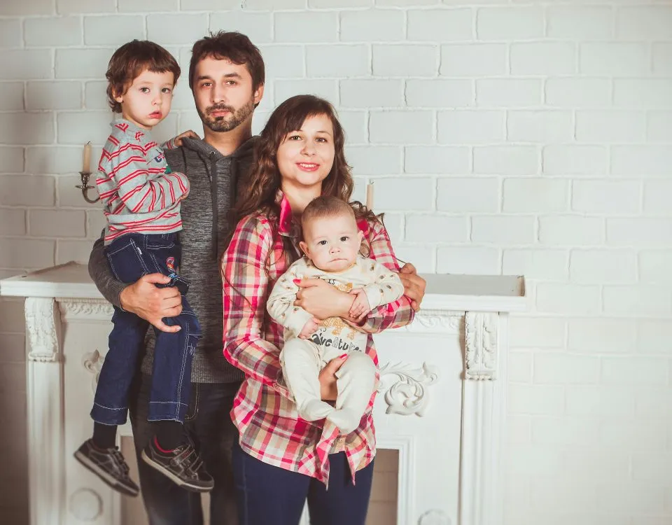A family poses in fron of a fireplace: 30-something dad holds a toddler and 30-something mom holds a baby in her arms.