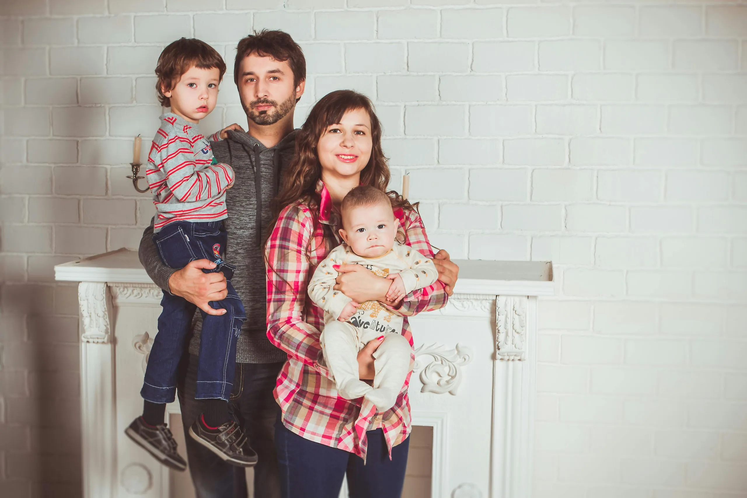 A family poses in fron of a fireplace: 30-something dad holds a toddler and 30-something mom holds a baby in her arms.
