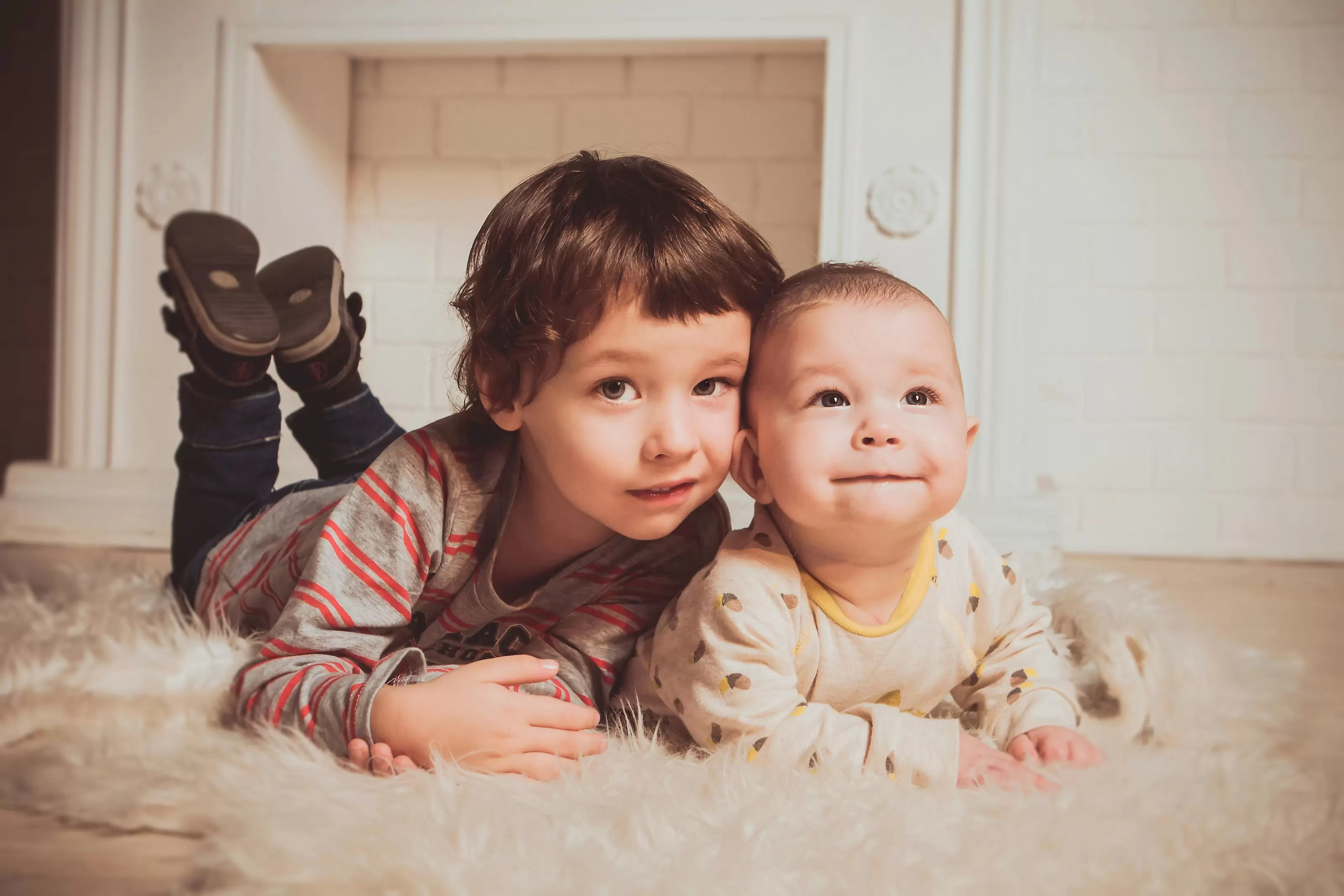 A toddler and baby in their pajamas lay on their bellies side by side on a fuzzy carpet, looking at the camera