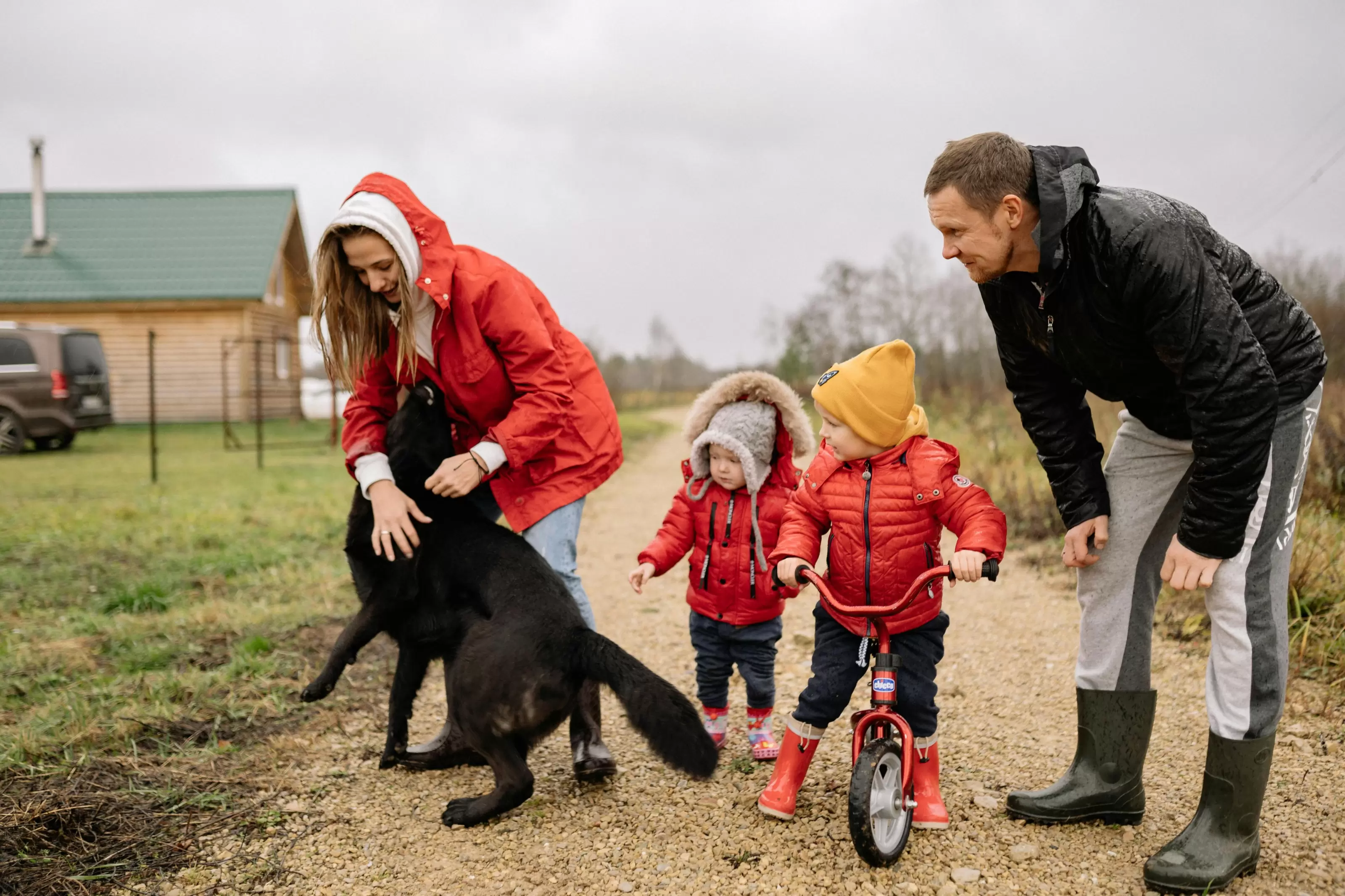 A family of 4 plays outside in their driveway. Dad stands on the right, leaning forward with hands on knees, next to a toddler on a bicycle and an infant standing behind them. they all watch as their mom plays with their large black dog.
