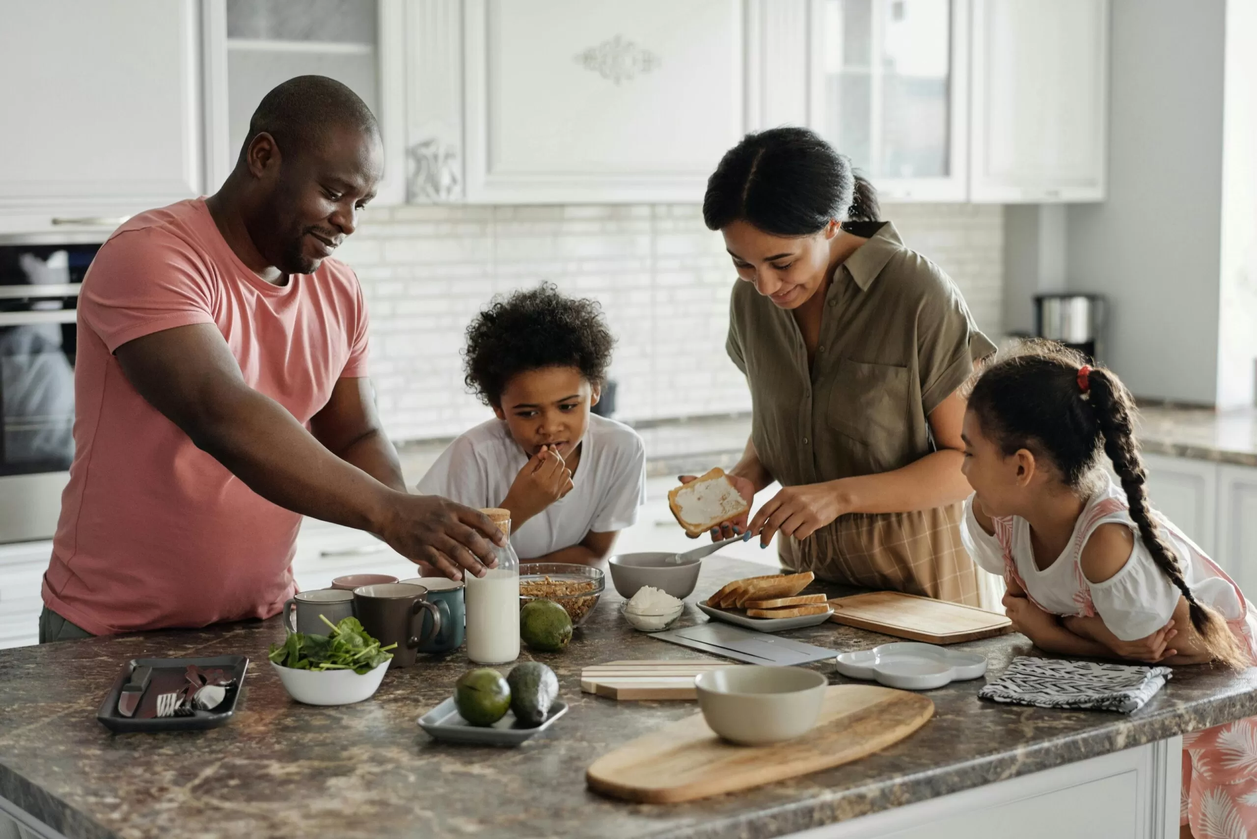 An African Canadian family gathers around a kitchen island preparing a breakfast: Dad sets the milk on the counter, mom butters toast, a young boy samples some granola and older sister looks on.
