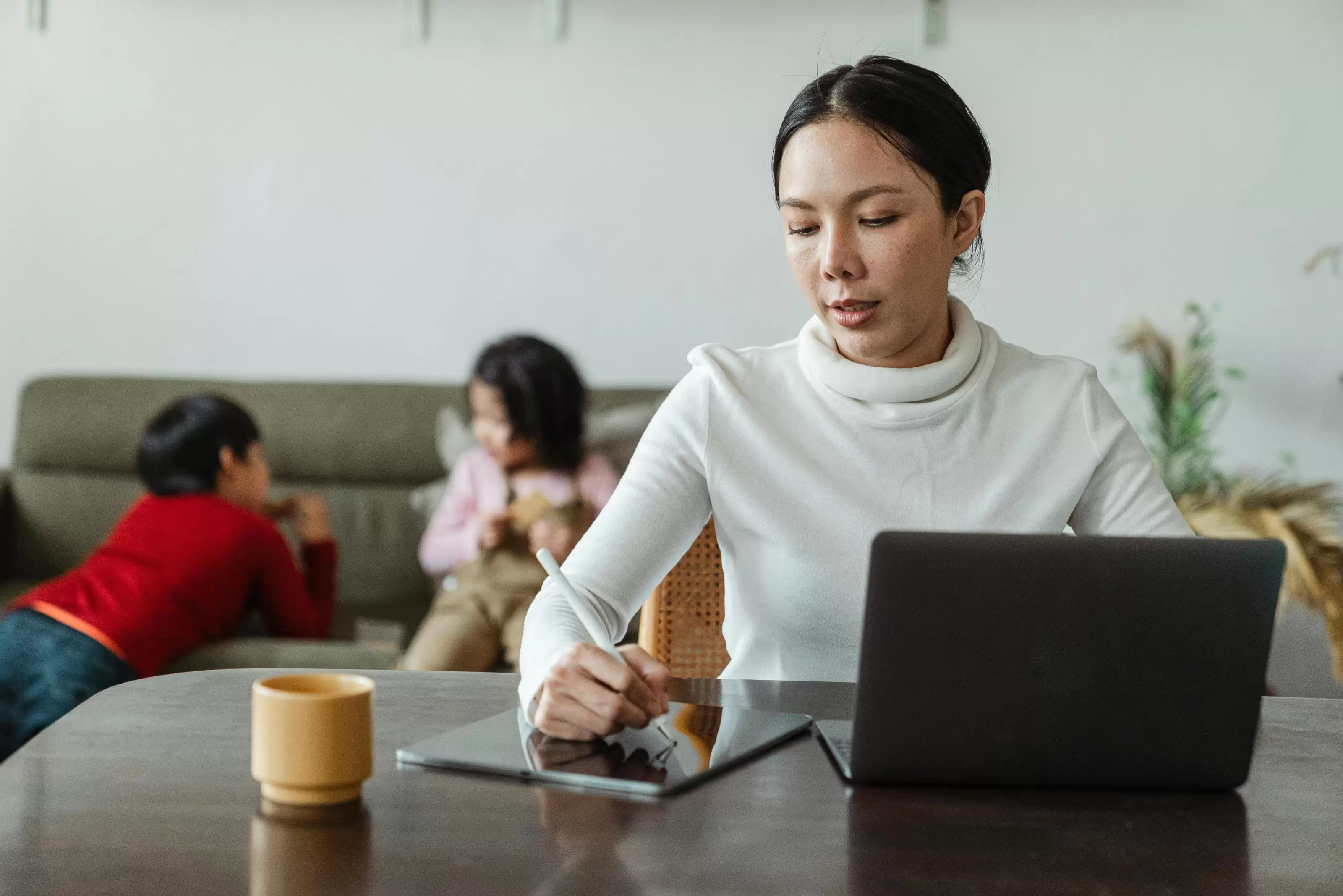 a woman sits at a table writing on a pad of paper with a laptop and cup of coffee in front of her. Two young kids play on the sofa behind her as she tries to work.