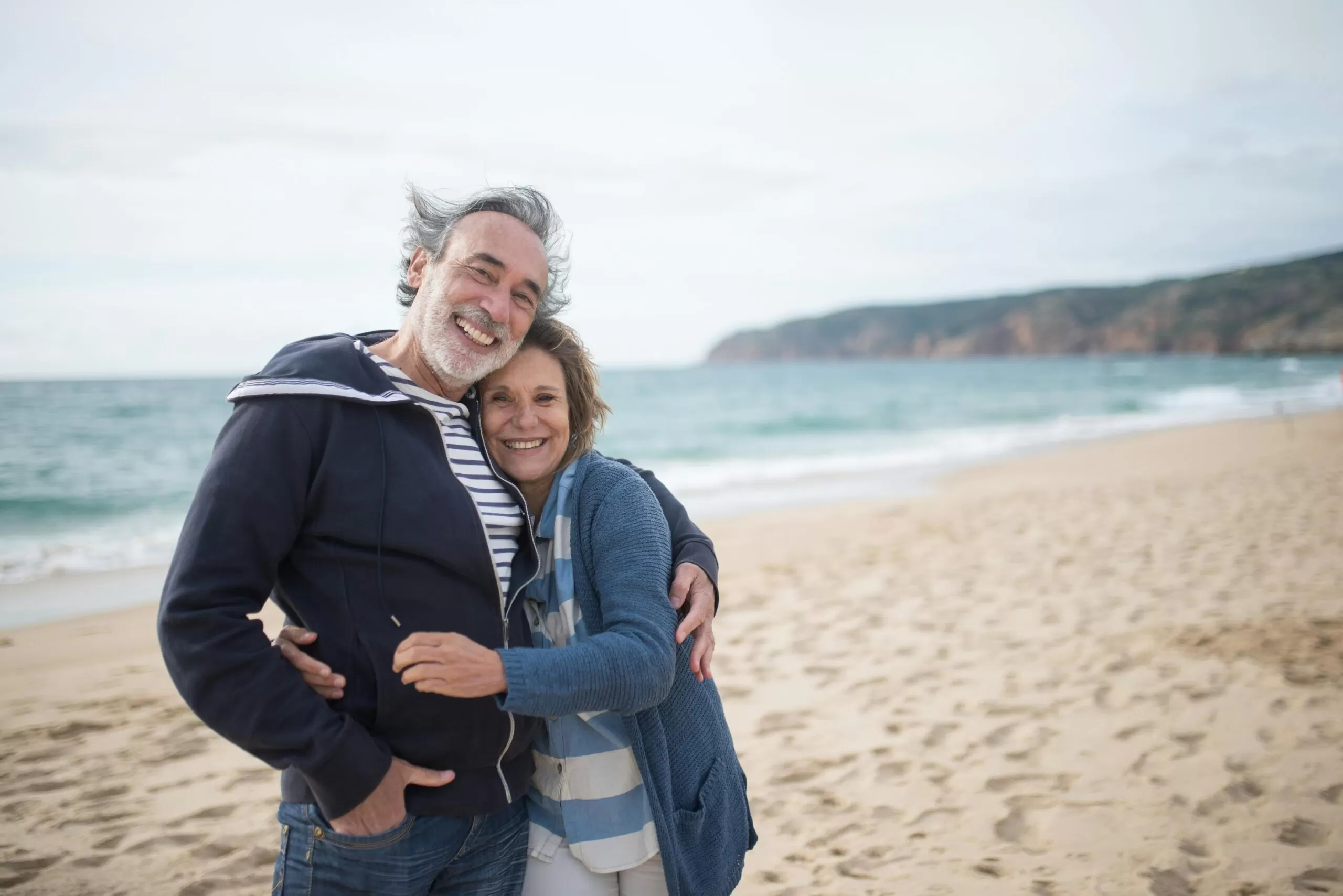 A senior couple hugs while standing on a beach, with the ocean and cliffs in the background