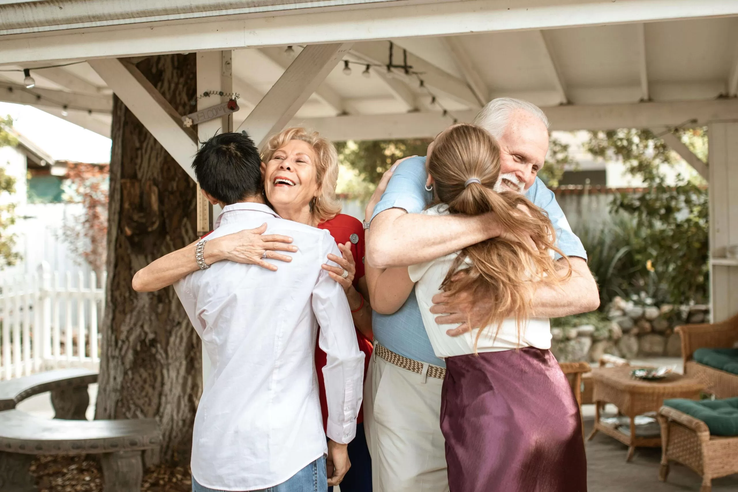 A senior woman smiles and hugs a young man in a white dress shirt and jeans, whose back is to the camera. Next to them a senior man with a white beard wearing a light blue shirt and khaki pants hugs a young woman with long hair. They are outside in a furnished pergola.