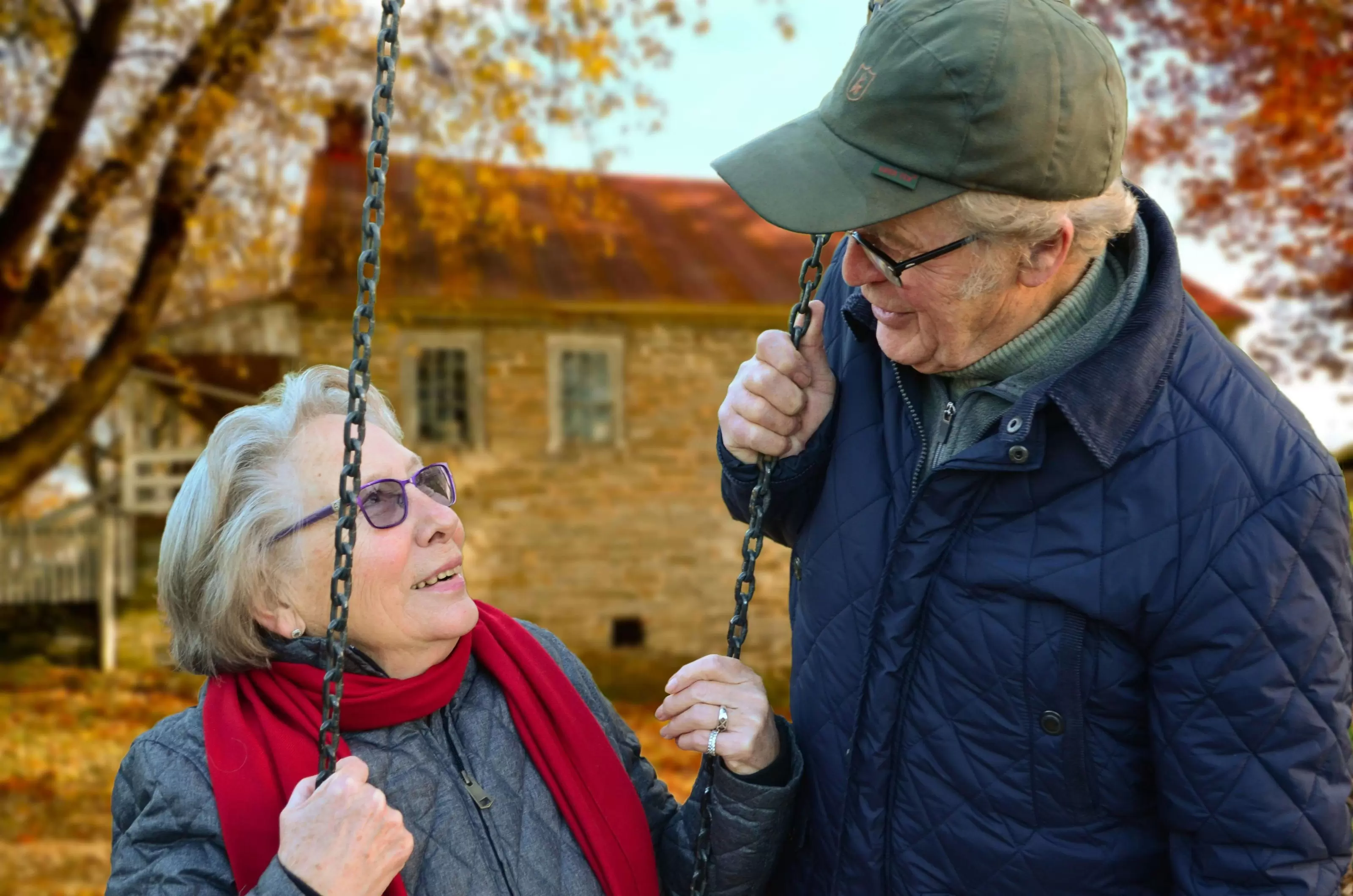 On an autumn day, a woman in her 70s wearing a jacket and red scarf sits on a swings and looks up at a man in his 70s wearing a baseball cap and jacket. He holds on of the chains of the swing and looks at her.
