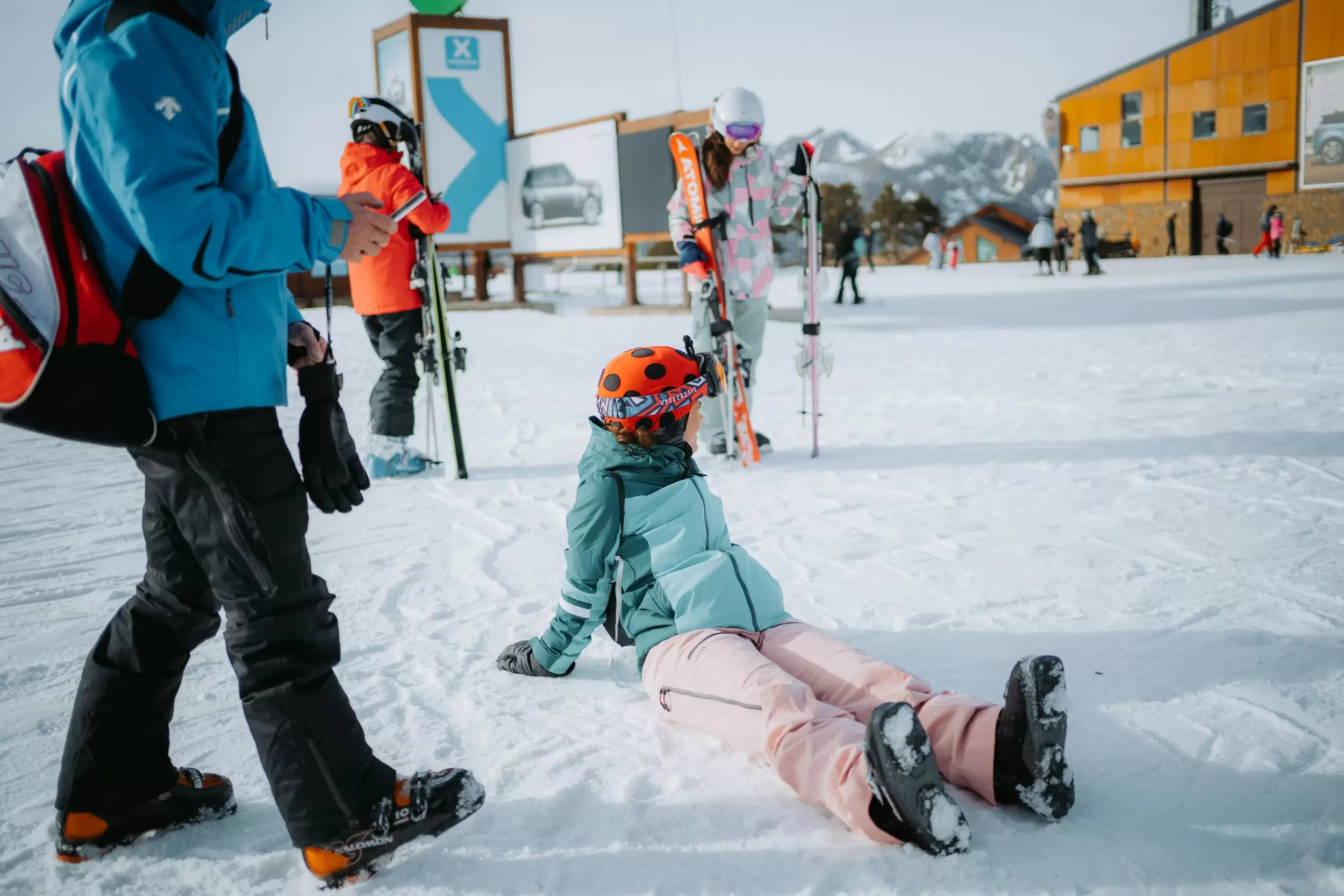 A child of about 10 years sits in the snow at a ski resort. Their legs are out in front of them, and they're resting back on their hands. They're wearing a downhill ski helmet with goggles sitting up on top of it, a ski jacket, snow pants, and ski boots. Another skier walks past looking at their phone. Other skiers are standing in the background. The child looks to the side at a building in the distance.