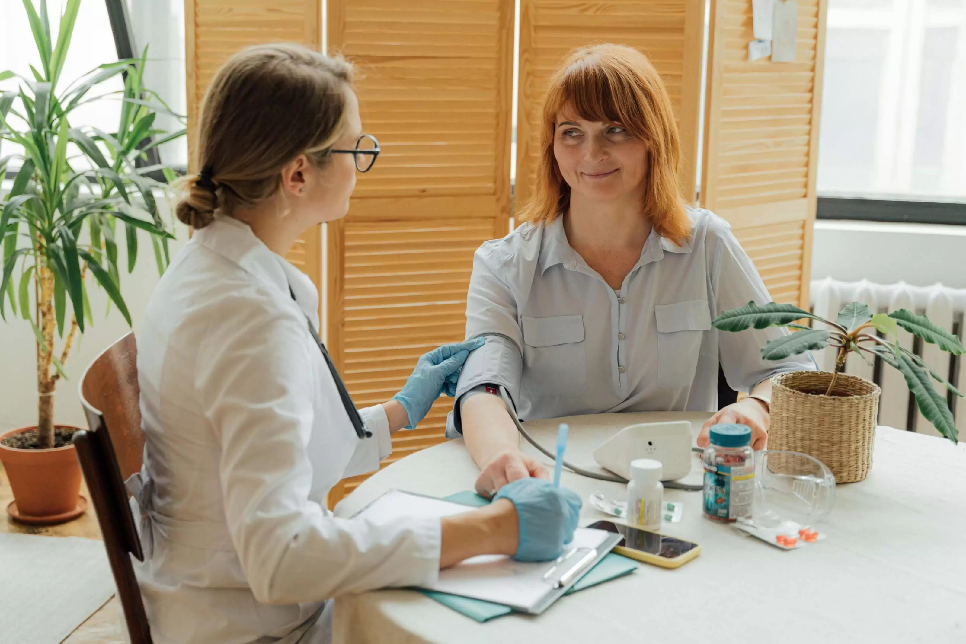 A female nurse wearing a lab coat and sterile gloves sits at a table in a kitchen setting. She is checking the blood pressure of a woman in her 30s who sits across from her, and writing on a medical chart. The woman looks calm and relaxed. On the table are a phone, 2 vitamin or medication bottles, and a potted plant.