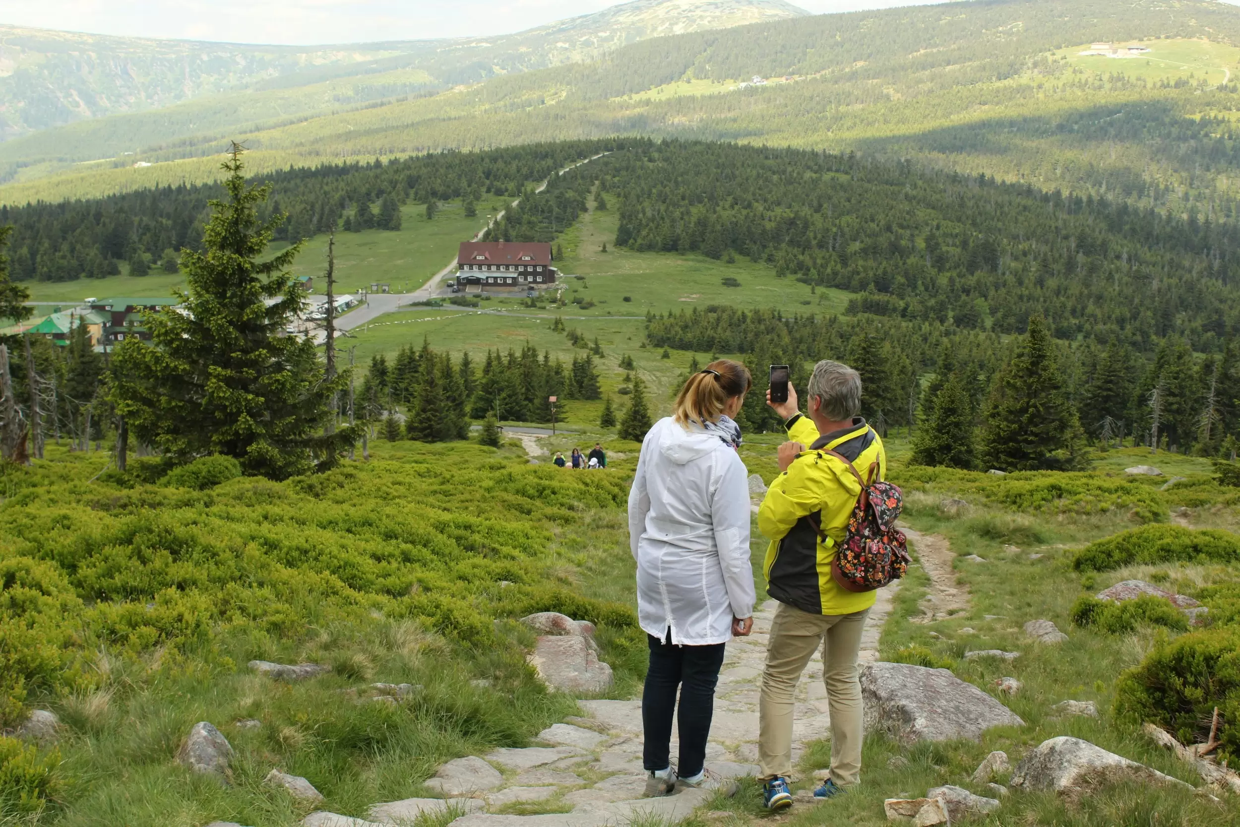 A mid-life couple in hiking clothes stand on the side of a mountain trail in Jelenia G&oacute;ra, Poland, taking a selfie.