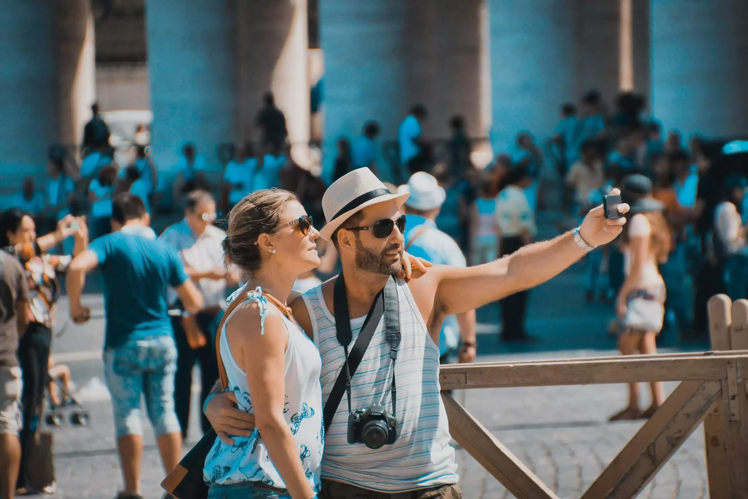 A tourist couple in their 20s stands in a public square taking a selfie. They are dressed for hot weather and sunshine.