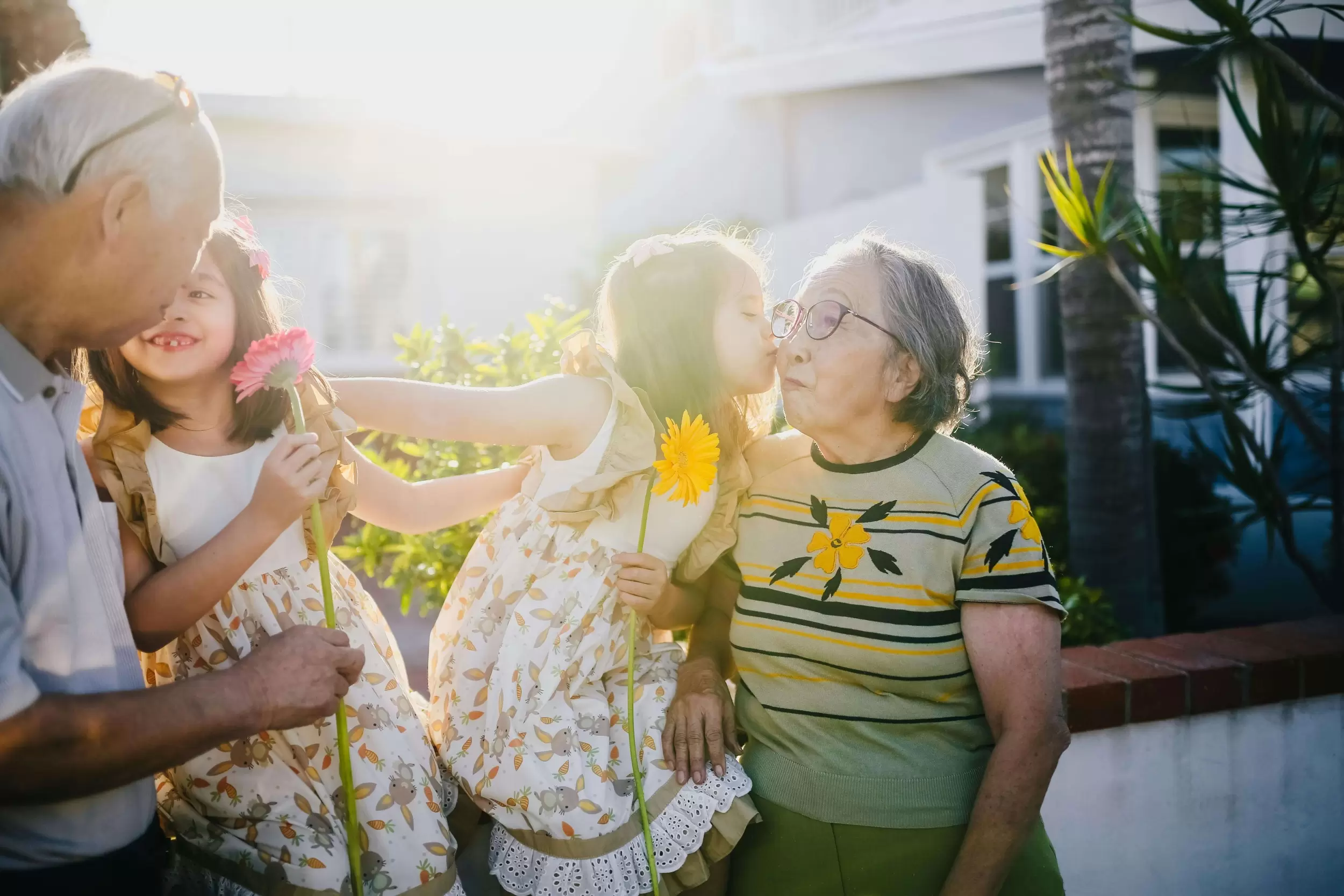 Two girls, about 7 years old, in matching sundresses and holding gerbera flowers are on a sunny deck with their grandparents. One kisses her grandmothers cheek, the other is laughing with her grandfather.