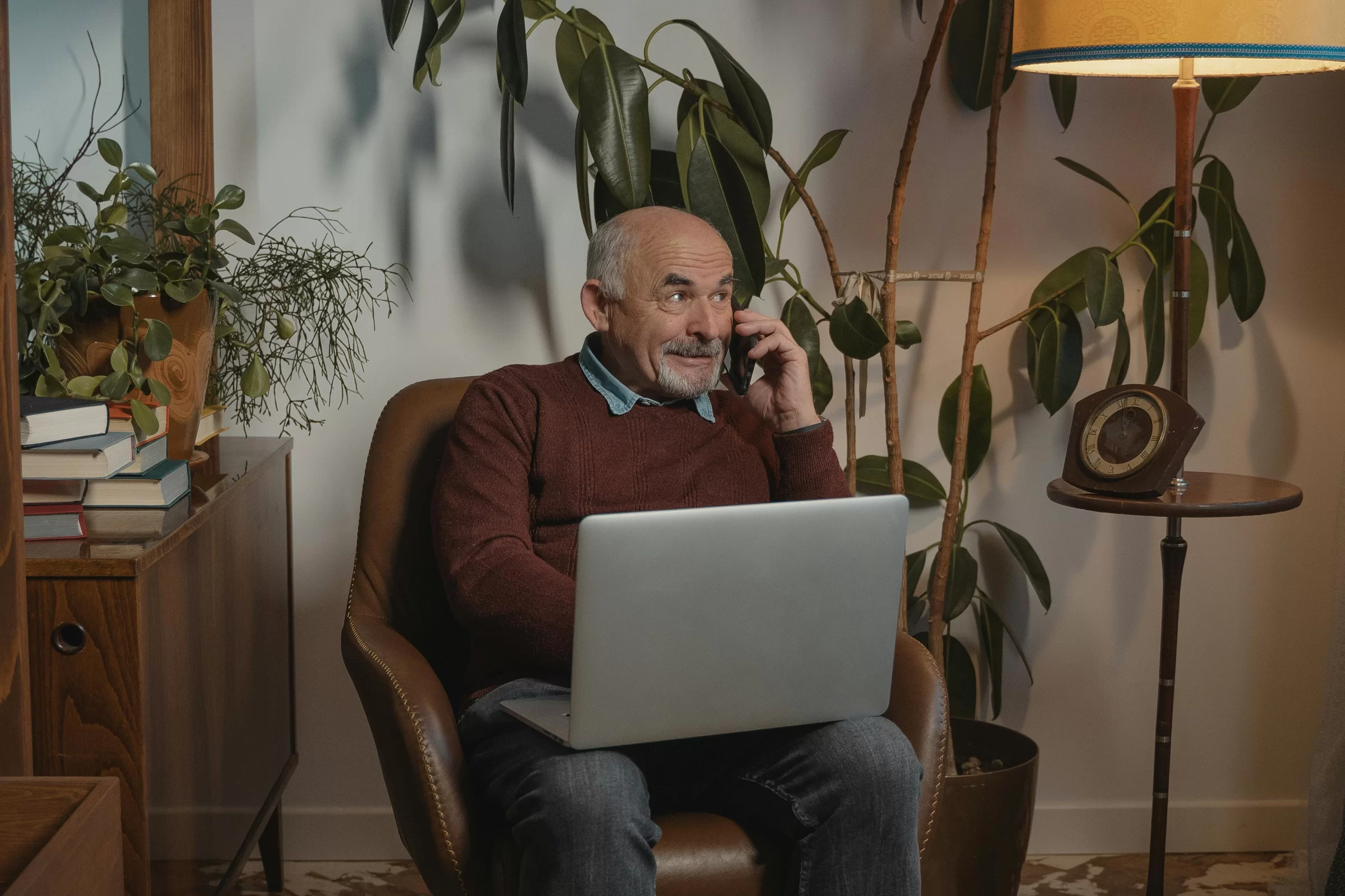 Senior man sitting in a chair in a living room talking on a cellphone, with a laptop on his lap. In the background are a tall plant, floor lamp, and credenza with books on it.