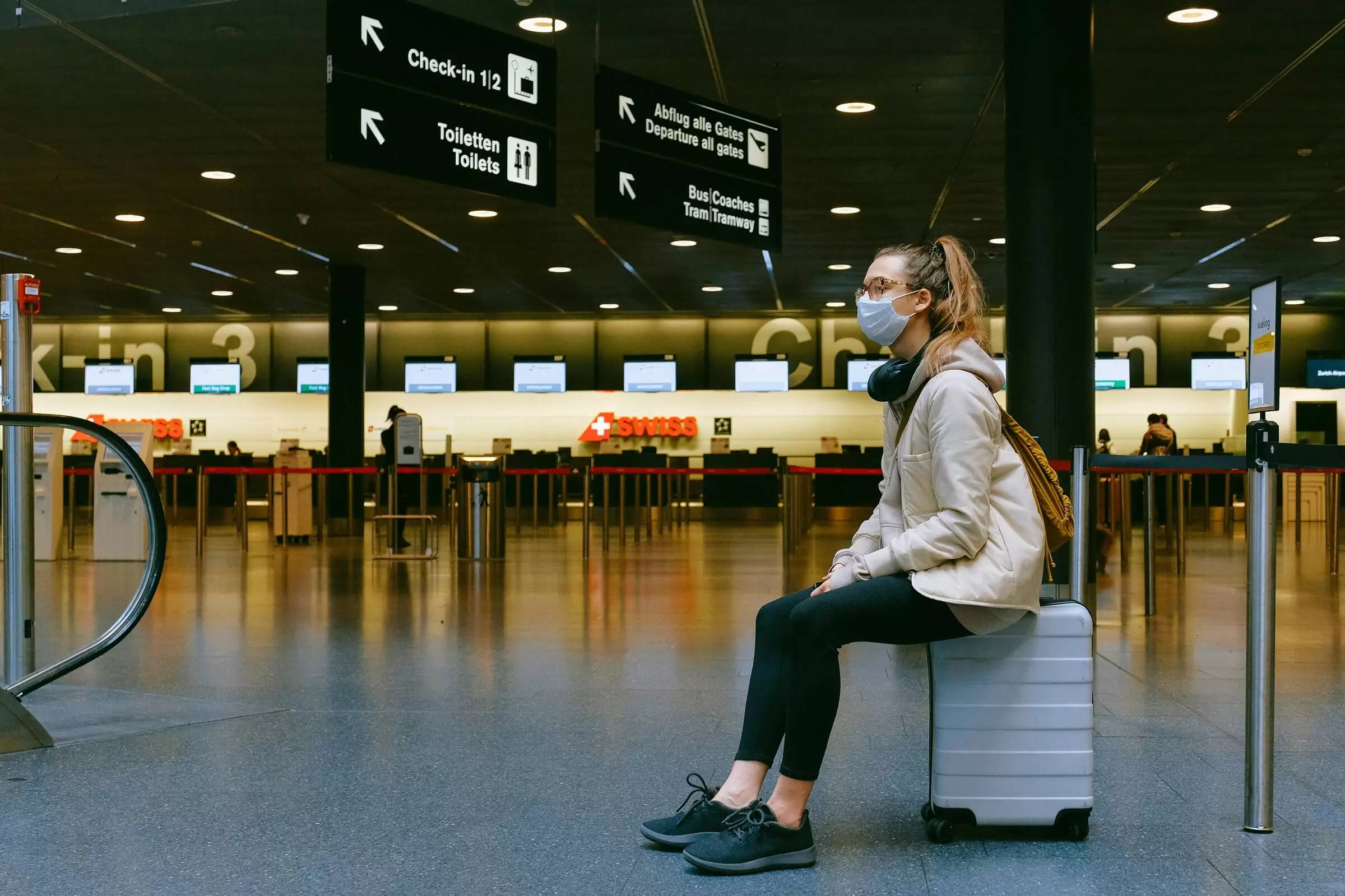 A young woman wearing a COVID mask and sits on her carry-on suitcase in the empty check-in area of a German airport.