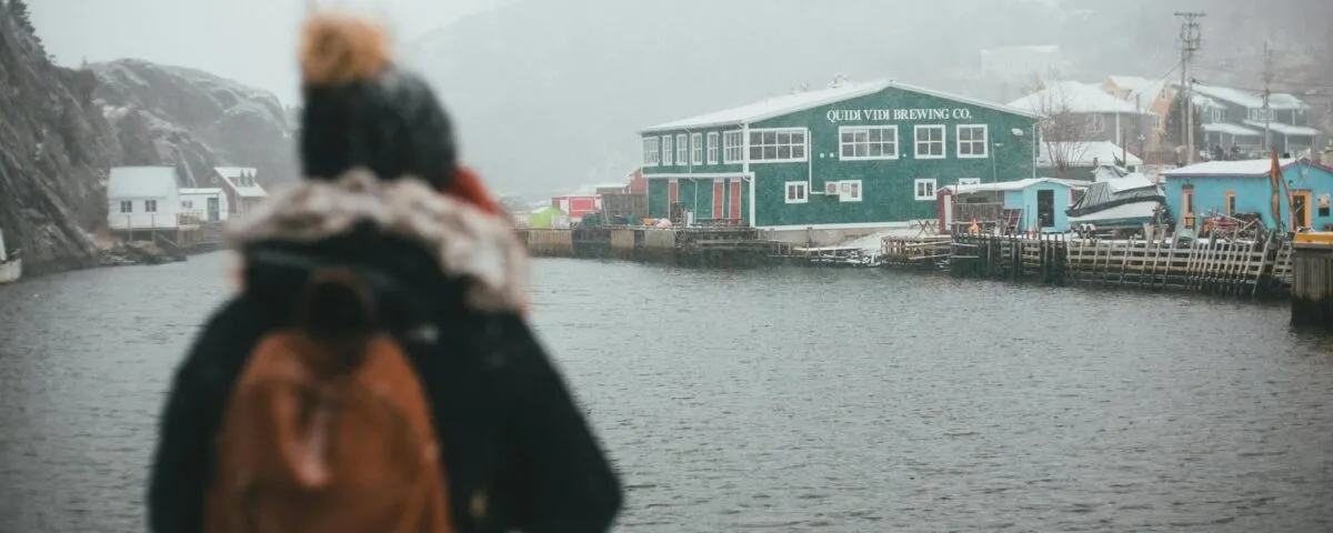A woman wearing a winter hat, jacket, and backpack stands by the harbour in St. John's Newfoundland looking at the Quidi Vidi Brewing Company building and coastline in the distance.