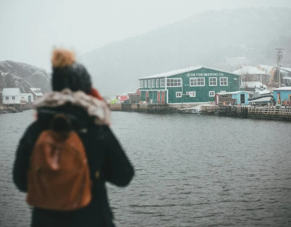 A woman wearing a winter hat, jacket, and backpack stands by the harbour in St. John's Newfoundland looking at the Quidi Vidi Brewing Company building and coastline in the distance.