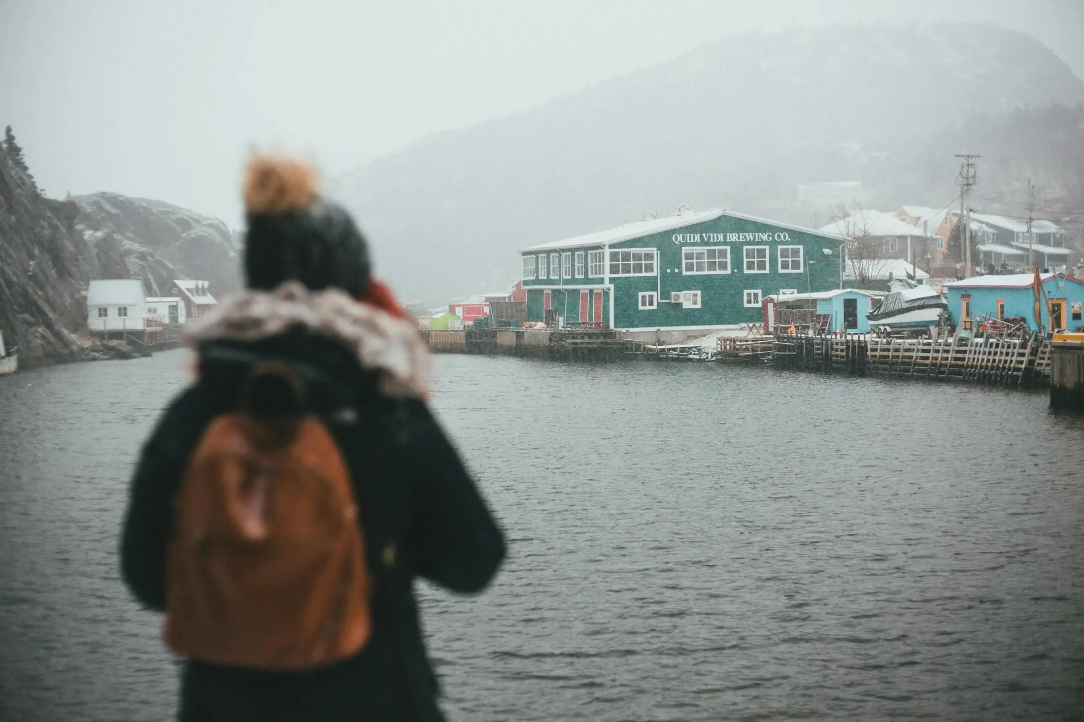 A woman wearing a winter hat, jacket, and backpack stands by the harbour in St. John's Newfoundland looking at the Quidi Vidi Brewing Company building and coastline in the distance.